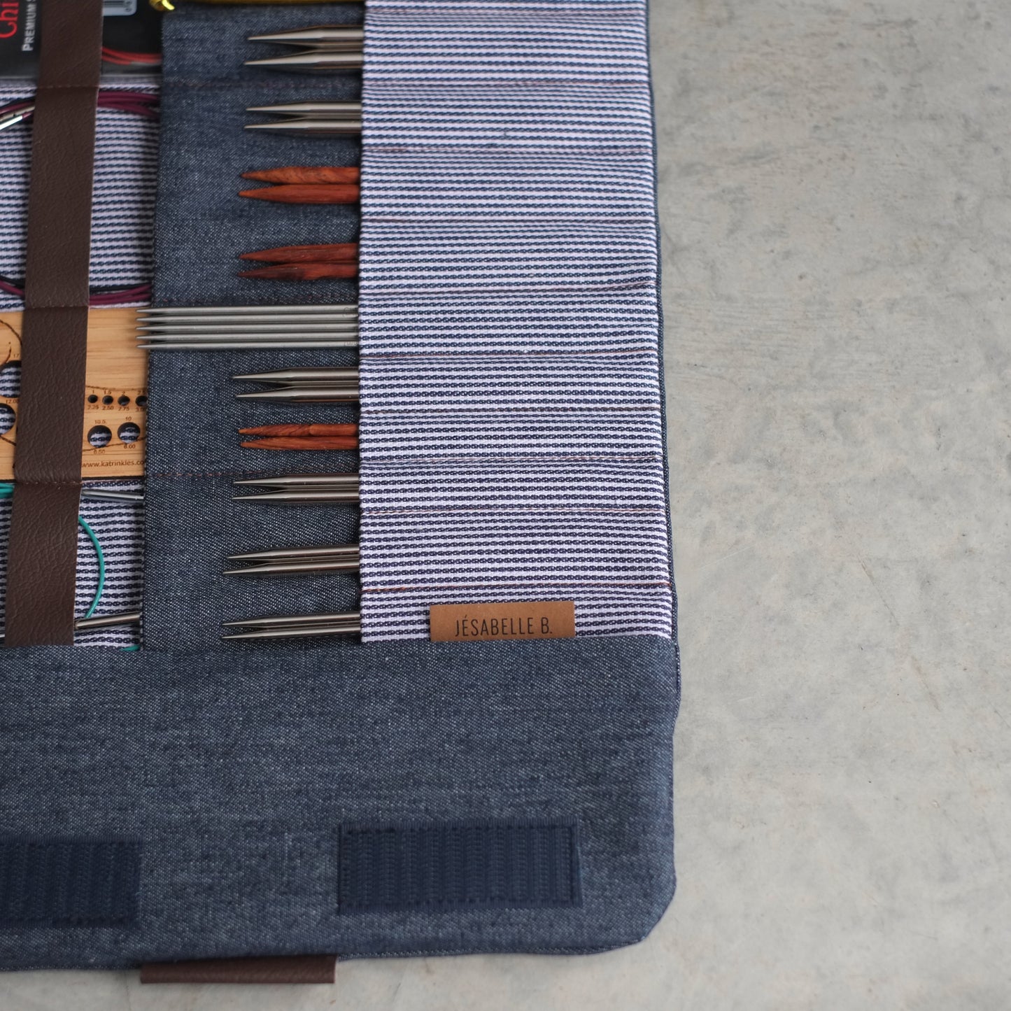 Gros plan sur le denim bleu foncé et l'étiquette Jésabelle B. — pointes d'aiguilles circulaires interchangeables bien rangées, fait main au Canada. | Close-up of dark denim fabric and Jésabelle B. label — interchangeable circular needle tips neatly stored, handmade in Canada.
