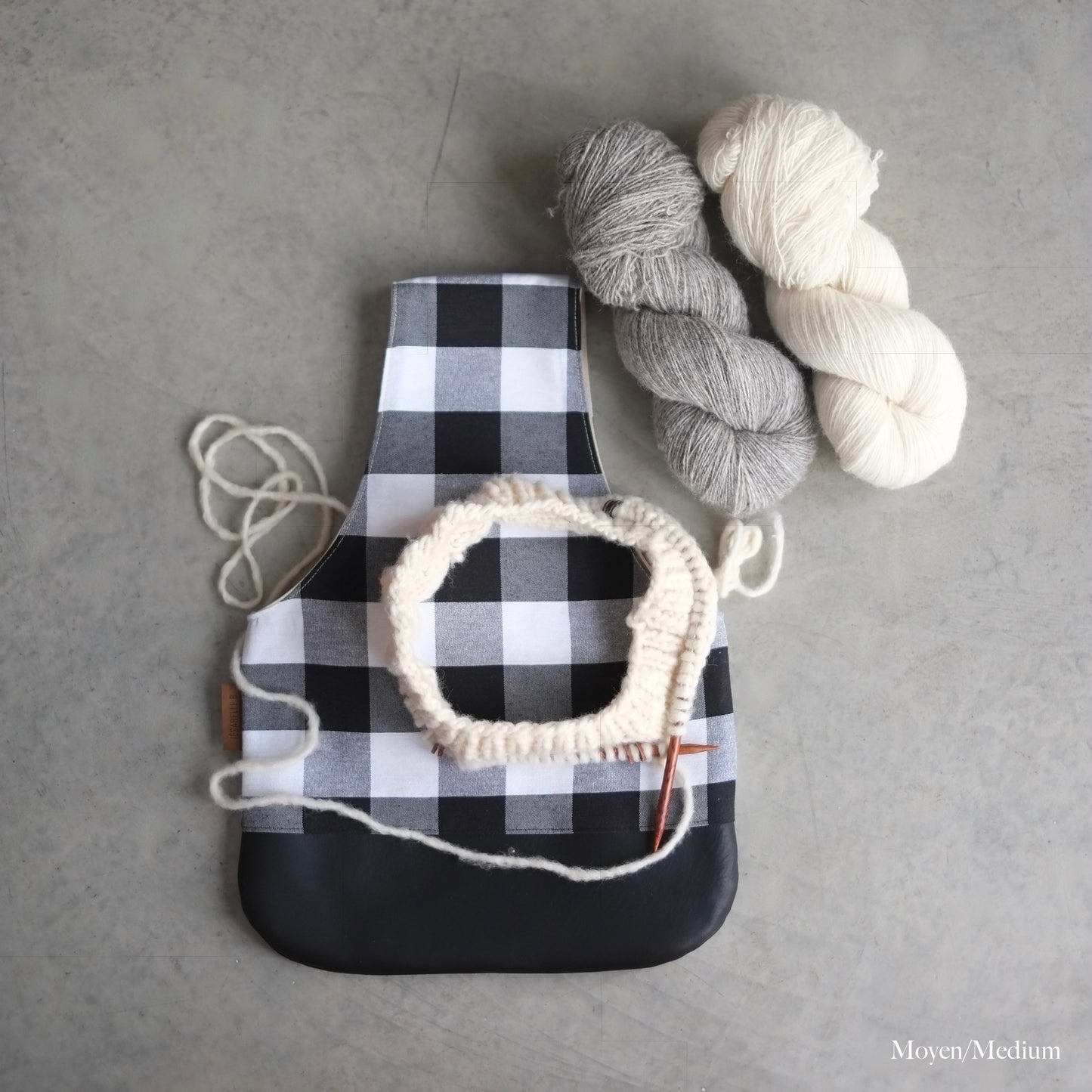 Top view of a black and white buffalo plaid knitting bag with a black leather bottom and yarn