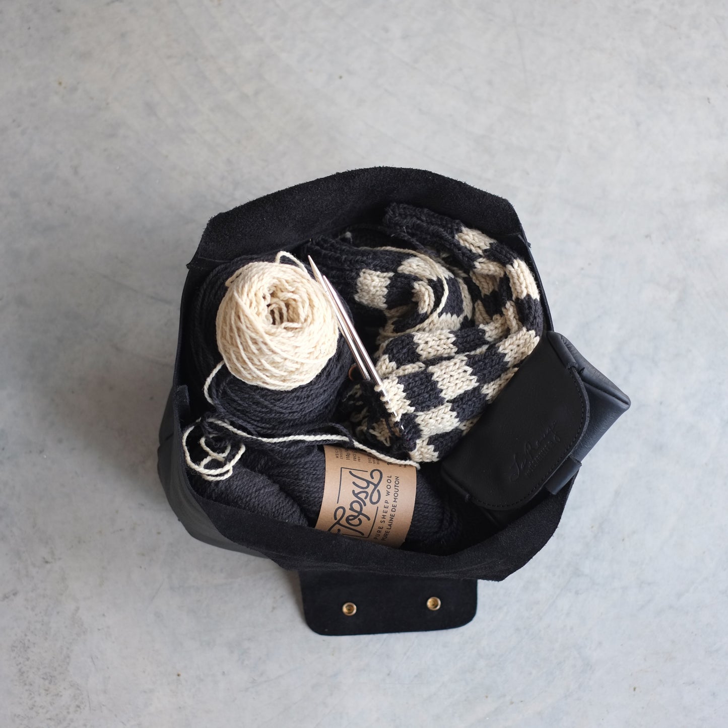 Top view of a large black leather bag full of yarn, needles and a case specific to a sweater project / Vue de haut d'un sac de projet ouvert où on voit clairement un projet chandail à l'intérieur avec la laine, les aiguilles et un étuid'Accessoires