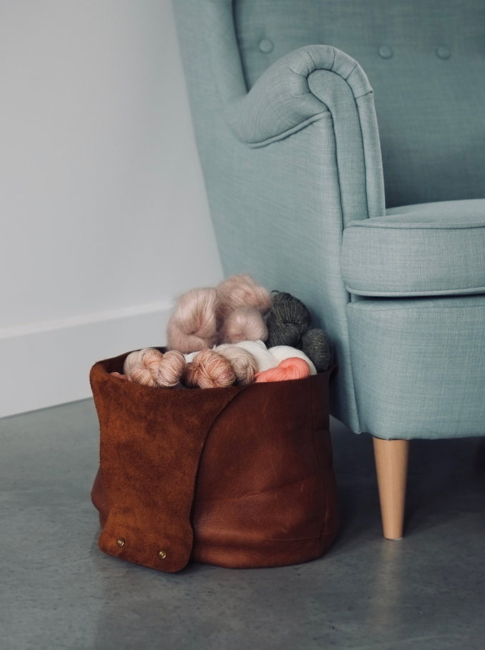 A large brown leather bag opened next to a sofa in a bucket position full of yarn / Un sac de tricot en cuir brun en position panier rempli de laine